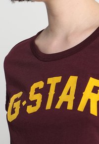 Camiseta color burdeos con cuello redondo y mangas cortas, que presenta un gran texto amarillo de "G-STAR" en el pecho. Tejido de algodón suave.