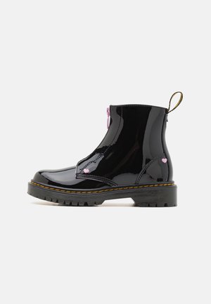 Bottine noire brillante à talon avec semelle épaisse à crampons et couture jaune, décorée de petits détails en forme de cœur rose et d'une languette arrière.