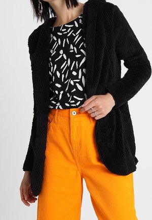 Cardigan negro y esponjoso abierto sobre una blusa de patrón en blanco y negro, combinado con pantalones naranjas de cintura alta. Presenta un corte holgado y mangas largas.
