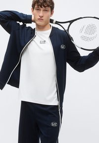 Tmavomodrá tepláková súprava s bielymi detailmi, obsahujúca bundu na zips s logom na hrudi a zodpovedajúce nohavice. Model drží tenisovú raketu.