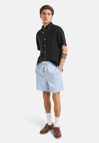 Kortermet svart lin-skjorte med lomme, sammen med lyseblå shorts med snor, hvite sokker og brune sko.