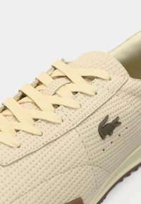 Μπεζ αεριζόμενα αθλητικά παπούτσια με κίτρινες κορδόνια, υφή φινιρίσματος και μπρονζέ λογότυπο κροκοδείλου. Διαθέτουν καφέ αντίθετη σόλα και επένδυση.