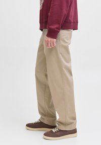 Pantalones beige de corte relajado, hechos de algodón con una textura suave, combinados con zapatillas marrón oscuro que tienen cordones blancos y una suela clara.