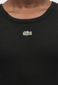 Κοντινό πλάνο ενός ατόμου που φοράει μαύρο πλεκτό πουκάμισο με ένα μικρό πράσινο λογότυπο κροκοδείλου στο κέντρο κάτω από το λαιμό.