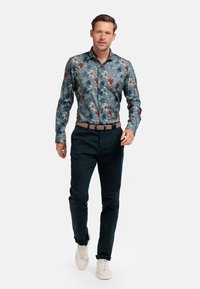Bloemmotief overhemden in teal, met rode, beige en groene accenten; gecombineerd met donkerblauwe broek en witte sneakers.