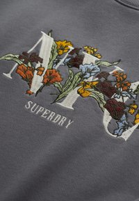 Sudadera gris con vibrantes flores bordadas en tonos rojos, naranjas, amarillos y azules, con el texto "Superdry" debajo.