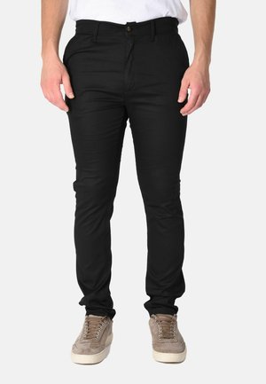 Uomo che indossa pantaloni neri slim fit e sneaker in camoscio marrone chiaro, in piedi davanti a uno sfondo semplice.