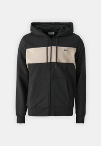 Haut à capuche zippé noir avec un panneau horizontal crème sur la poitrine, comportant un petit logo vert. Tissu doux avec poches avant.
