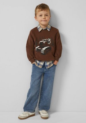 Brauner Pullover mit Traktor-Graphic, über einem blauen karierten Hemd getragen; kombiniert mit weit geschnittenen blauen Jeans und weißen Sneakern.