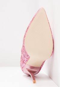 Chaussure à talon haut pointu pailletée rose avec un talon stiletto fin, posée sur le côté contre une surface et un mur blancs.