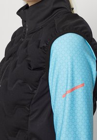 Person som bär en svart quiltad väst över en ljusblå mönstrad långärmad skjorta med en liten röd textetikett på ärmen.