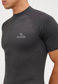 Korte mouw rash guard in donkergrijs, gemaakt van rekbare stof. Bevat flatlock naden en een klein wit logo op de borst.