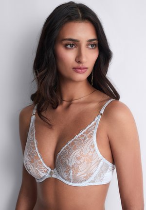 Femme aux longs cheveux foncés portant un soutien-gorge en dentelle blanche transparente, regardant légèrement vers la droite sur un fond clair uni.