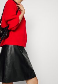 Personne portant un pull en tricot rouge et une jupe en cuir noire, portant un petit sac noir à l'épaule, vue de profil sur un fond uni.