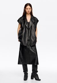 Melna ādas pārāk liela veste ar plašiem atlokiem un divu pogu aizdari, apvienota ar garu melnu ādas svārku un pieguļošiem melniem zābakiem.