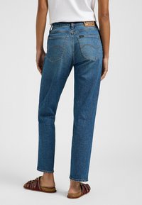 Jeans in denim blu con taglio dritto, caratterizzati da cuciture arancioni, tasche posteriori e un'etichetta color sabbia in vita. Sandali con elementi decorativi.