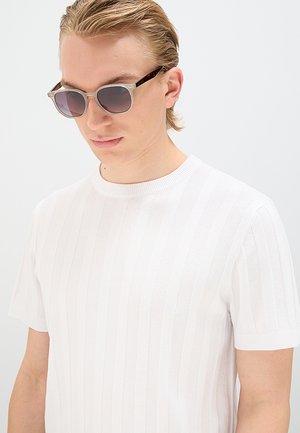 Giovane uomo con capelli biondi corti che indossa occhiali da sole e una maglia bianca a maniche corte lavorata a trama, su sfondo semplice.