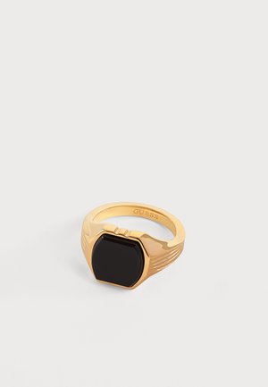 Bague chevalière en or avec une pierre hexagonale noire polie et un anneau strié, gravée du nom de la marque à l'intérieur.