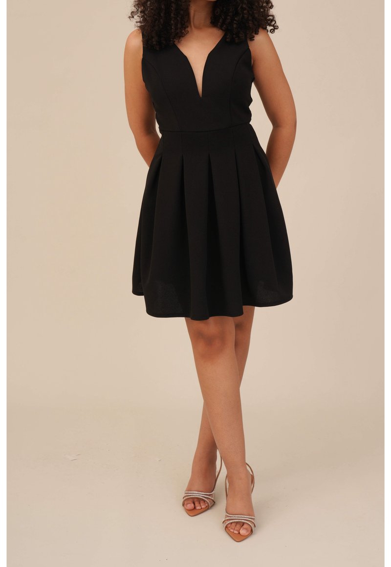 Femme portant une robe noire sans manches avec un décolleté en V profond et une jupe plissée, associée à des sandales à talons beiges.