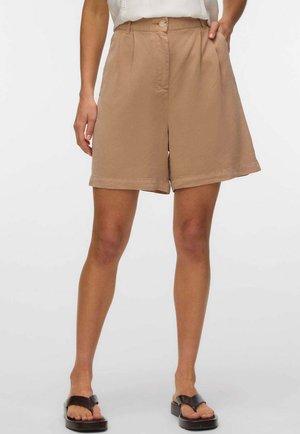 Donna che indossa pantaloncini larghi color sabbia con vita alta, bottone e zip frontali, abbinati a sandali neri con punta aperta e top bianco.