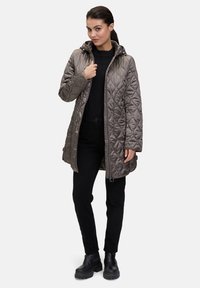 Pikowana, długa szara kurtka z kapturem, z zapięciem na zamek i kieszeniami po bokach. Nosi w zestawie z czarną bluzką i czarnymi spodniami oraz masywnymi butami.