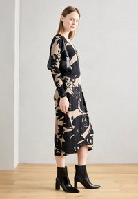 Musta kukkakuosiset mekko päällä mallilla, jossa on muotoon ommeltu yläosa ja levenevä hame, yhdistettynä mustiin nilkkabootseihin ja pieneen mustaan laukkuun.