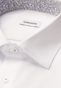Chemise blanche avec un col intérieur fleuri et une étiquette cousue portant la mention "seidensticker" près de l'encolure.