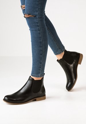 Botas Chelsea de cuero negro con paneles laterales de elastano, puntera redondeada y tacón bajo marrón, usadas con jeans ajustados azules desgastados.