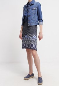 Giacca di jeans con chiusura a bottoni, colore blu; gonna a metà coscia a fantasia in blu navy con design geometrici; scarpe blu senza lacci.