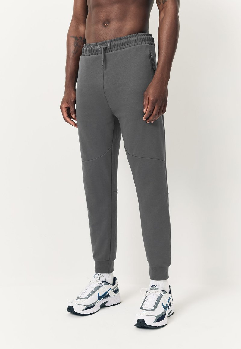 Gray jogger pants with a drawstring waistband, side zipper pockets, and stitched panel details. Worn with white and navy athletic shoes.
