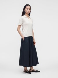 Femme aux longs cheveux foncés portant un polo blanc à manches courtes, une longue jupe noire avec des poches et des ballerines slingback noires avec des boucles.