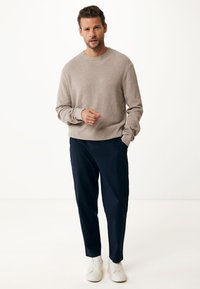 Helle beige Strickjacke mit Rundhalsausschnitt, kombiniert mit dunkelblauen Hosen. Das Model trägt weiße Sneaker; die Hände entspannt an den Seiten. Schlichtes Design.