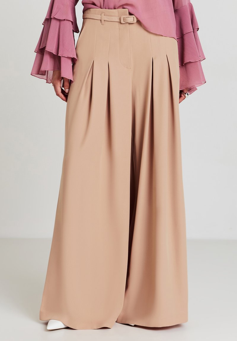 Pantaloni beige a vita alta e gamba larga con pieghe frontali, una cintura abbinata e una texture del tessuto liscia, abbinati a una blusa rosa con maniche a volant.