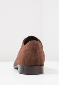 Chaussure en daim marron avec un bout arrondi, un profil bas et un talon en bois sombre. La texture est douce et mate, avec des détails de couture minimes.