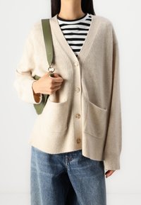Beżowy wełniany kardigan z dekoltem w serek, prążkowanymi mankietami i dwoma przednimi kieszeniami, w zestawieniu z czarno-białą koszulą w paski i dżinsami.