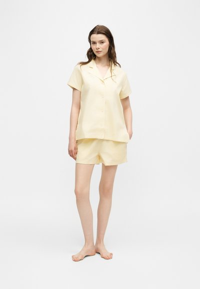 Femme debout pieds nus portant une chemise boutonnée à manches courtes jaune pâle et un short assorti sur un fond clair uni.