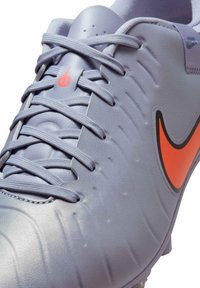 Chaussure de football synthétique grise avec un dessus texturé, un swoosh Nike orange et des lacets gris ; présente des lignes surélevées sur la surface.