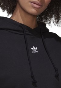 Svart hoodie med dragsko på huvan och en vit Adidas-logga på bröstet. Materialet verkar mjukt och har en texturerad yta.