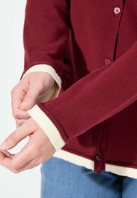 Bordeaux cardigan med vita manschetter och fåll, med knappar, ribbad textur och rund halsringning. Händer justerar ärmarnas manschetter.