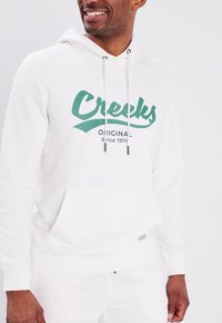 Sudadera blanca con capucha y bolsillo canguro, con un logo verde de "Creeks" y el texto "Original Desde 1974". Tejido suave de mezcla de algodón.