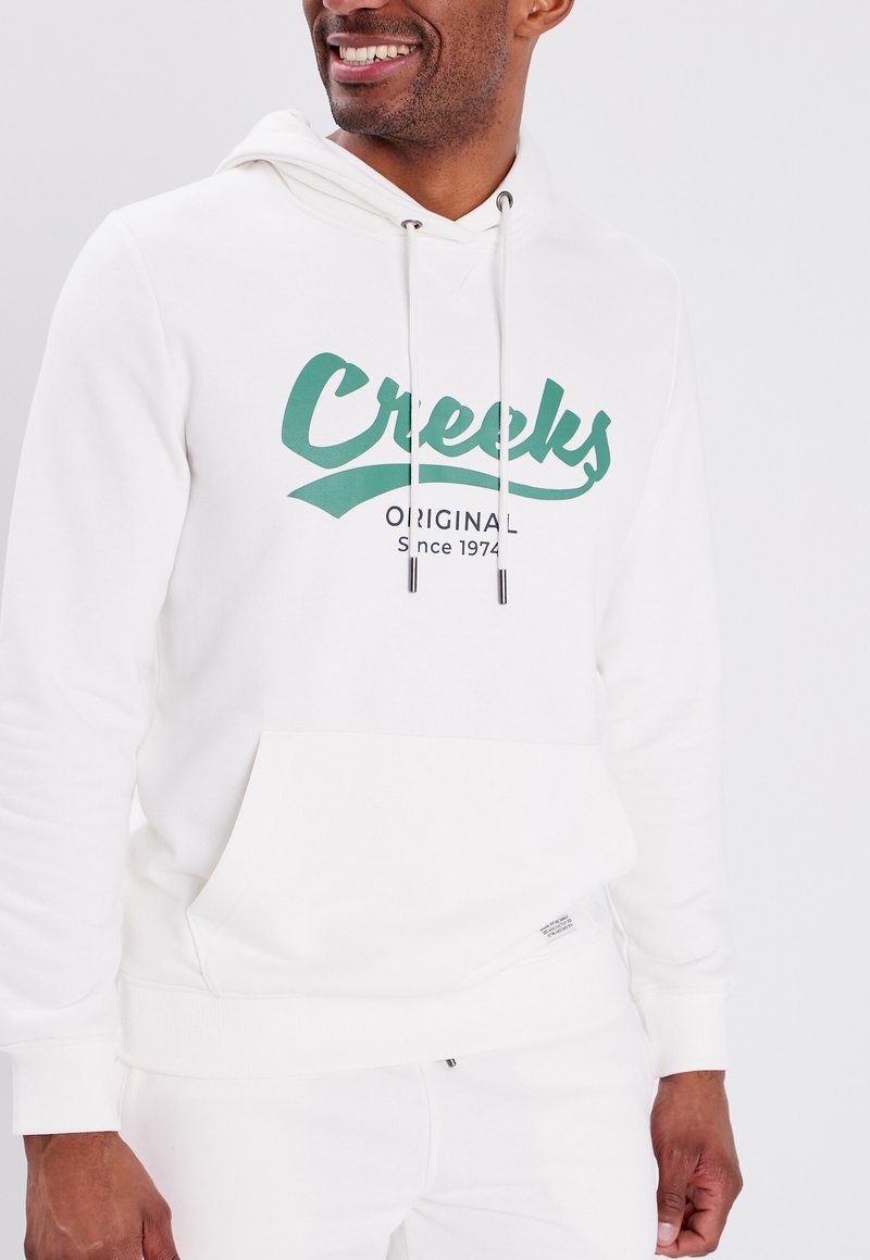 Sudadera blanca con capucha y bolsillo canguro, con un logo verde de "Creeks" y el texto "Original Desde 1974". Tejido suave de mezcla de algodón.