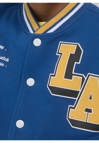 Niebieska kurtka varsity z białymi zatrzaskami, ściągaczem przy kołnierzu z żółto-białymi paskami oraz dużym napisem "LA" w kolorze żółtym i czarnym.