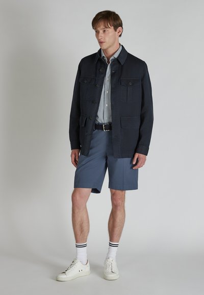 Giacca navy con tasche, indossata sopra una camicia a righe blu chiaro. Pantaloni corti grigi e sneakers bianche con strisce nere sui calzini completano l'outfit.