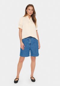 Lichte crèmekleurige blouse met knoopsluiting en korte pofmouwen, gecombineerd met blauwe denim shorts. Zwarte instappers maken de outfit compleet. Eenvoudig en casual ontwerp.