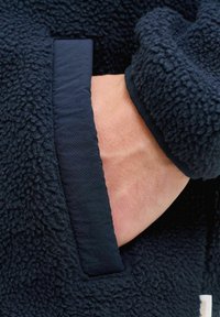 Mână introdusă în buzunarul unei jachete fleece de culoare albastru închis, cu material texturat și margine netedă a buzunarului.