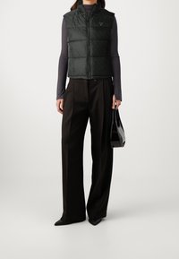Gilet trapuntato verde scuro con collo alto, chiusura con zip e texture a motivo. Abbinato a pantaloni neri a gamba larga e una elegante borsa nera.