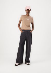 Brown cotton t-shirt with embroidered design, paired with wide-leg gray pants. The model wears a pink cap and sneakers.