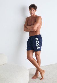 Jūriniai maudymosi šortai su baltais "BOSS" logotipais, raišteliu juosmens srityje, lengvos medžiagos ir laisvos silueto, derinami su nuogomis kojomis ir neutralia fono spalva.