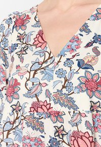 Blumenmusterstoff mit V-Ausschnitt, der pinke und blaue Blumen, grüne Blätter und braune Äste auf einem cremefarbenen Hintergrund zeigt.