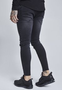 Svarta smala jeans med en slät yta, med bakfickor och en figurnära design, tillsammans med svarta sportskor.